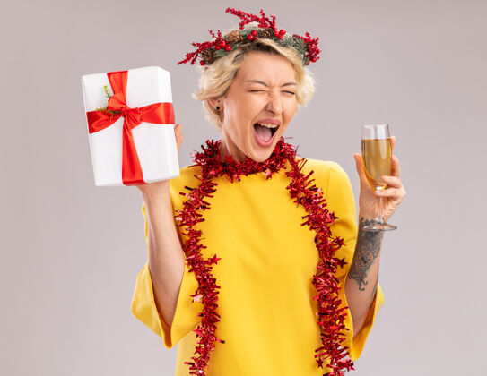 年轻快乐的金发女郎戴着圣诞花环 脖子上戴着金属丝花环 手里拿着香槟和礼包 闭着眼睛在白色背景上尖叫周围圣诞节礼物