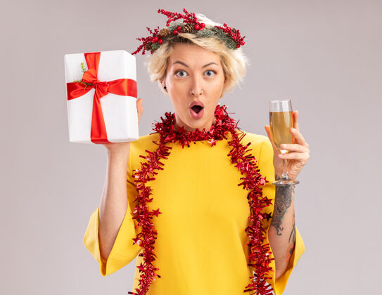 香槟年轻的金发女郎戴着圣诞花环 脖子上戴着金箔花环 手里拿着香槟和礼包 看着隔离在白色背景上的相机圣诞年轻白色
