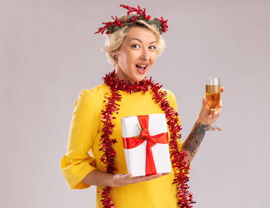 香槟年轻的金发女郎戴着圣诞花环 脖子上戴着金箔花环 手里拿着香槟和礼包 看着隔离在白色背景上的相机圣诞玻璃女人