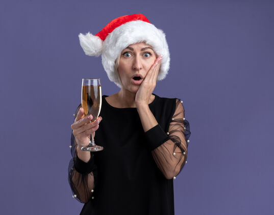 复制一个戴着圣诞帽的中年金发女人 手举香槟 看着镜头 手放在脸上 在紫色的背景下与世隔绝金发关心圣诞快乐