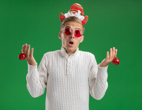 抱印象深刻的年轻帅哥戴着圣诞老人的头带戴着眼镜拿着圣诞装饰球看着隔离在绿色背景上的相机圣诞快乐圣诞老人小伙子