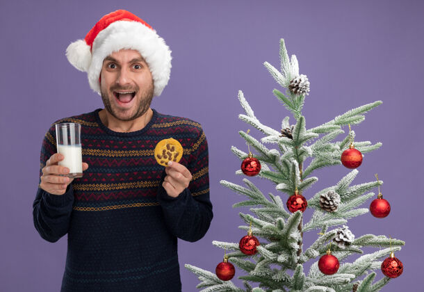 帽子令人印象深刻的年轻白种人戴着圣诞帽站在装饰圣诞树附近 拿着一杯牛奶和饼干 看着紫色背景上孤立的摄像机年轻人玻璃牛奶