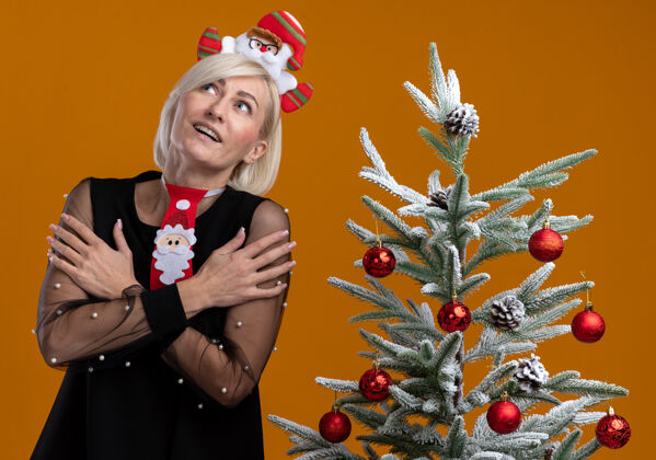 手臂令人印象深刻的中年金发女人戴着圣诞老人的头带和领带站在装饰圣诞树附近双手交叉在手臂上看着橙色背景孤立圣诞老人印象中年