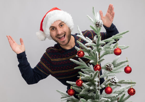 印象令人印象深刻的年轻白人男子戴着圣诞帽站在圣诞树后看着相机保持双手在空气中隔离在白色背景圣诞树白年轻