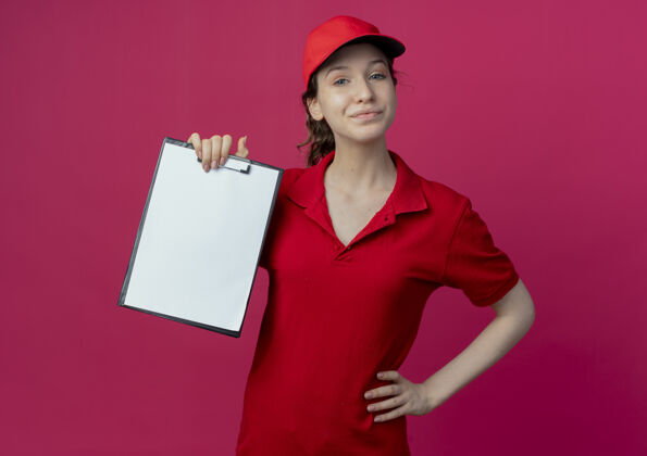 深红色高兴的年轻漂亮的送货女孩在红色制服和帽子显示剪贴板和把手放在腰部隔离在深红色的背景帽子手年轻
