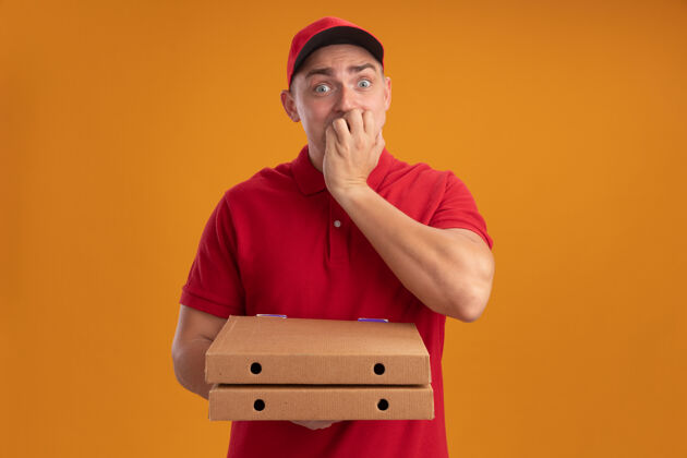 人关心的年轻送货员穿着制服 戴着帽子 拿着披萨盒 手放在嘴巴上 隔离在橙色的墙上公民年轻人站着