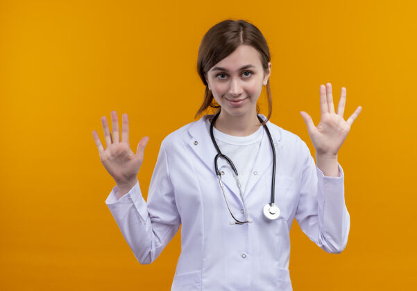 空虚自信的年轻女医生穿着医用长袍和听诊器 在孤立的橙色空间里展示空手自信年轻听诊器