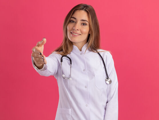 脸年轻的女医生穿着医用长袍 手持听诊器 站在隔离在粉红色墙上的门前感觉穿着年轻人