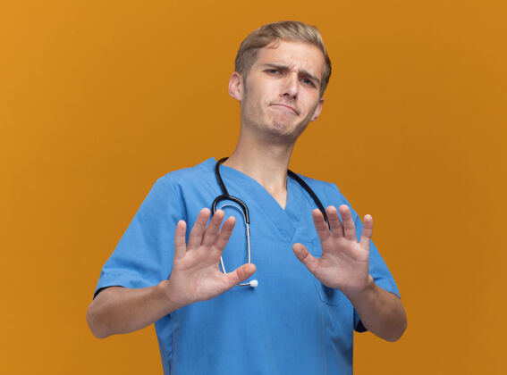 站着身穿医生制服 手持听诊器的年轻男医生 手伸向隔离在橙色墙上的摄像机人脸表情