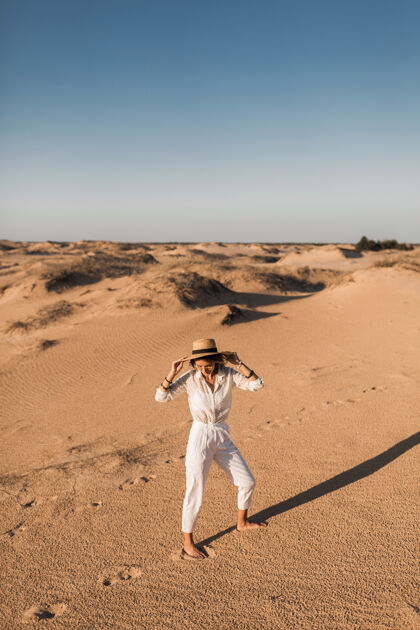 人时尚美丽无忧无虑的快乐女人走在沙漠沙滩上穿着白色裤子和衬衫 戴着草帽在日落波西米亚夏天冒险
