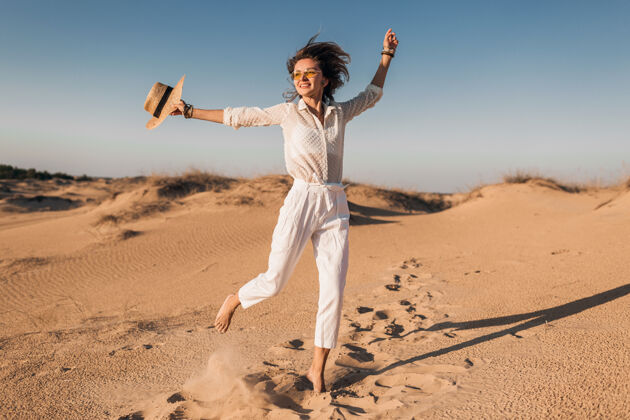自然时尚美丽的微笑开朗的女人在沙漠里快乐地奔跑着 穿着白色的裤子和衬衫 戴着草帽在夕阳下夏天旅游人