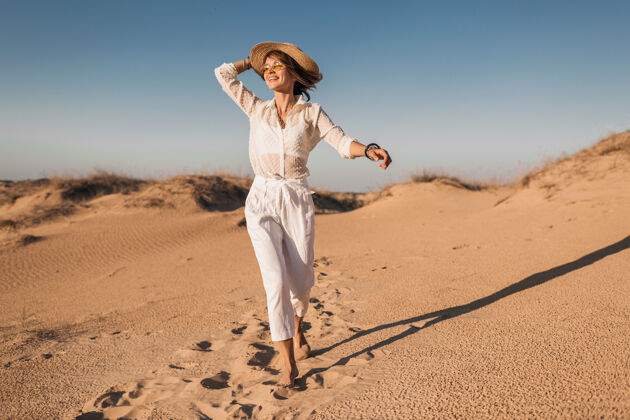 帽子时髦的微笑美丽的快乐女人穿着白色的衣服在沙漠中奔跑和跳跃在日落时戴着草帽女士快乐衬衫