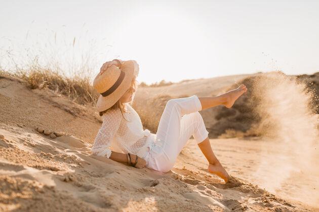 配饰落日时分 穿着白色衣服 戴着草帽和太阳镜 在沙漠中摆出一副时髦 快乐 迷人 微笑的女人造型时尚旅游时尚