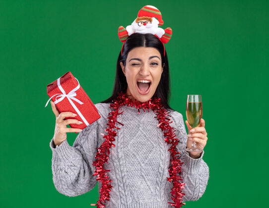 圣诞快乐快乐的年轻白人女孩戴着圣诞老人的头带 脖子上戴着金属丝的花环 手里拿着礼包和一杯香槟 看着相机在绿色背景上眨眼金属丝香槟礼物