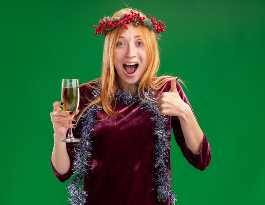 拇指兴奋的年轻漂亮女孩身穿红色连衣裙 脖子上戴着花环和花环 手持香槟酒杯 在绿色背景上孤立地竖起大拇指新年快乐花圈圣诞快乐