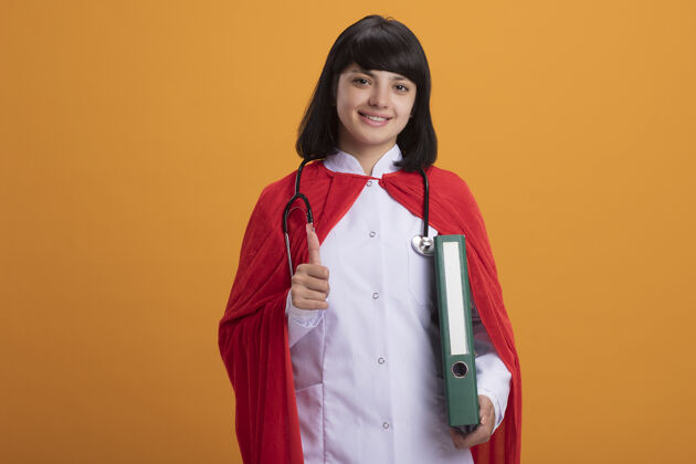 向上微笑着的年轻超级英雄女孩戴着听诊器 穿着医用长袍和斗篷 拿着文件夹 竖起大拇指微笑长袍年轻