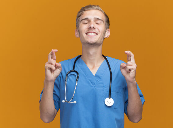 表情高兴地闭上眼睛年轻的男医生穿着医生制服听诊器交叉手指隔离在橙色的墙上年轻人闭合听诊器