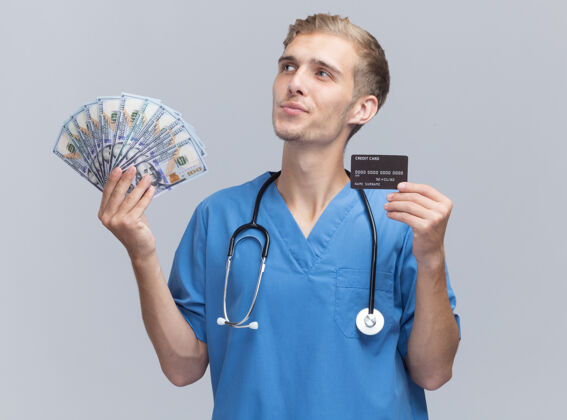 男性令人印象深刻的看边年轻的男医生穿着医生制服听诊器拿着现金和信用卡隔离在白色的墙上持有信用感情