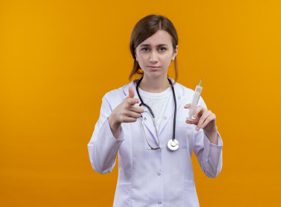 拿着自信的年轻女医生穿着医用长袍和听诊器拿着注射器 在孤立的橙色空间和复印空间做你的手势手势医疗医生