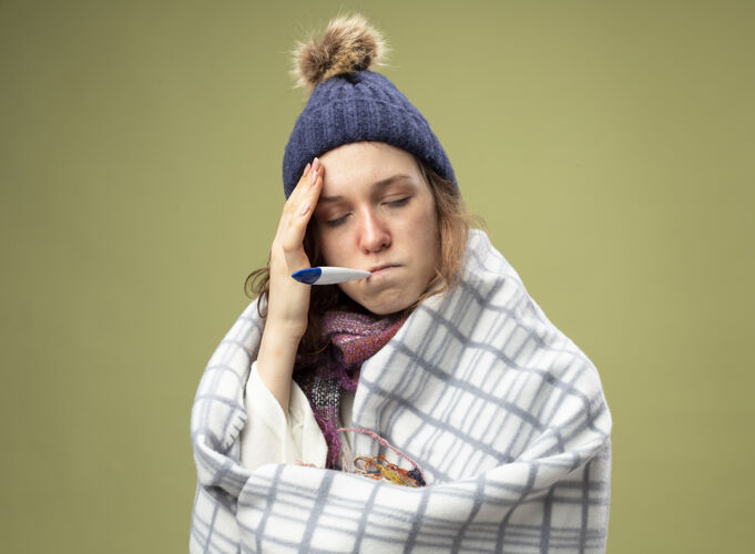 眼睛弱小的生病的小女孩 闭着眼睛 穿着白色长袍 戴着冬天的帽子 围巾用格子布包着 把温度计放在嘴里 把手放在额头上 隔离在橄榄绿上裹橄榄色年轻