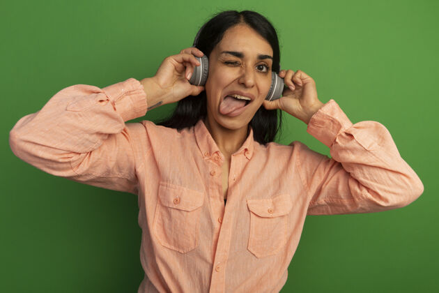 女孩眨眼的年轻漂亮女孩穿着粉色t恤 头戴耳机 舌头被隔离在绿色的墙上年轻舌头戴
