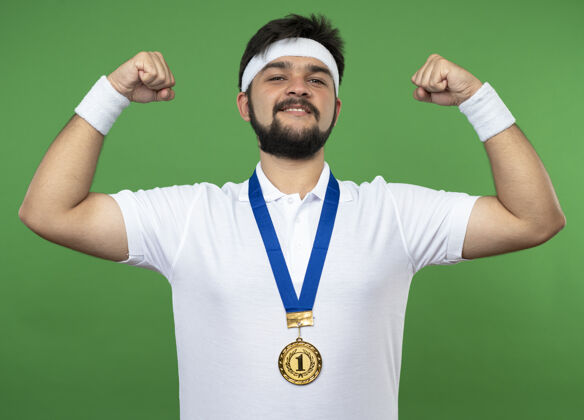 强壮微笑的年轻人戴着头带和腕带与奖牌显示出强烈的姿态隔离在绿色运动男子穿腕带