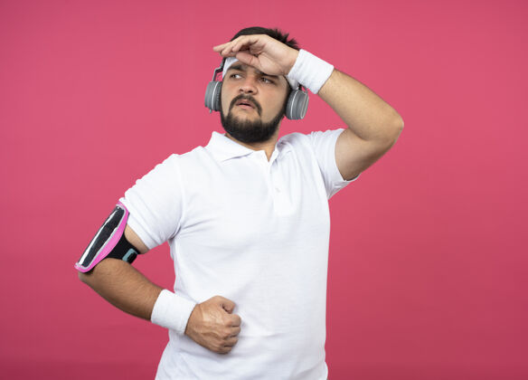 乐队自信的年轻运动型男子戴着头带和腕带 戴着耳机和电话臂带 手放在隔离在粉色墙上的前额上手臂手男人