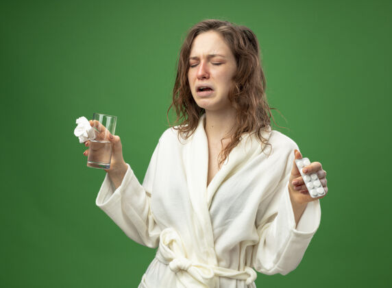 持有打喷嚏的生病的年轻女孩穿着白色长袍 手里拿着一杯水 把药片隔离在绿色上年轻长袍绿色