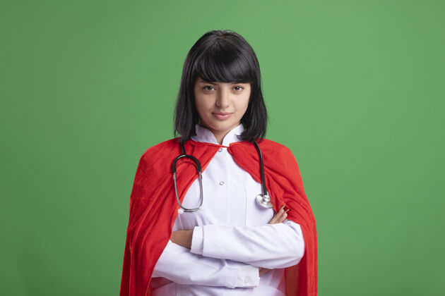 听诊器自信的年轻超级英雄女孩 戴着听诊器 穿着医用长袍和斗篷 双手交叉 隔离在绿色地带穿超级英雄斗篷