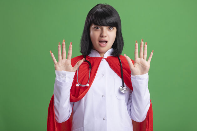 医疗惊讶的年轻超级英雄女孩戴着听诊器和医疗袍和斗篷展开双手隔离在绿色蔓延穿手
