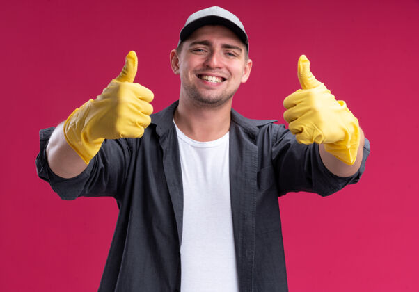 年轻面带微笑的年轻汉姆森清洁工穿着t恤和帽子 戴着手套 在粉红色的墙上孤立地竖起大拇指手套帽子清洁