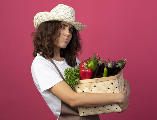 帽子悲伤的年轻女园丁穿着制服戴着园艺帽拿着菜篮子孤立在粉红色制服园艺女性