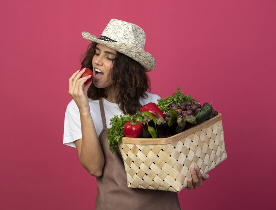 试试高兴的年轻女园丁穿着制服 戴着园艺帽 拿着菜篮子 试着把西红柿隔离在粉色上花园女性园艺