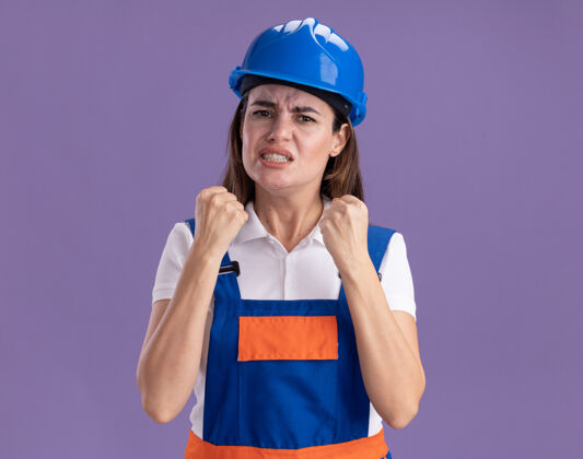 年轻愤怒的穿着制服的年轻建筑工人们在紫色的墙上孤立地握着拳头拳头握着建筑工人