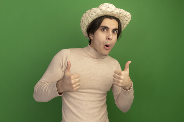 年轻年轻帅哥戴着帽子 大拇指竖立在绿色的墙上 看上去很高兴穿帅哥拜托