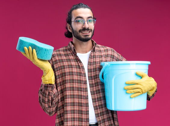 水桶年轻帅气的清洁工穿着t恤 戴着手套 手里拿着一桶海绵 隔离在粉红色的墙上帅哥戴着年轻人
