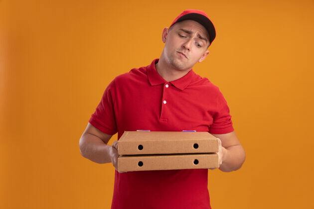 困惑困惑的年轻送货员穿着制服 戴着帽子 看着隔离在橙色墙上的比萨饼盒制服拿着披萨