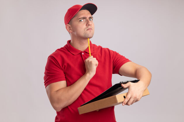 男人严格的年轻送货员穿着制服戴着帽子拿着剪贴板拿着披萨盒把铅笔放在下巴上隔离在白色的墙上剪贴板严格年轻人