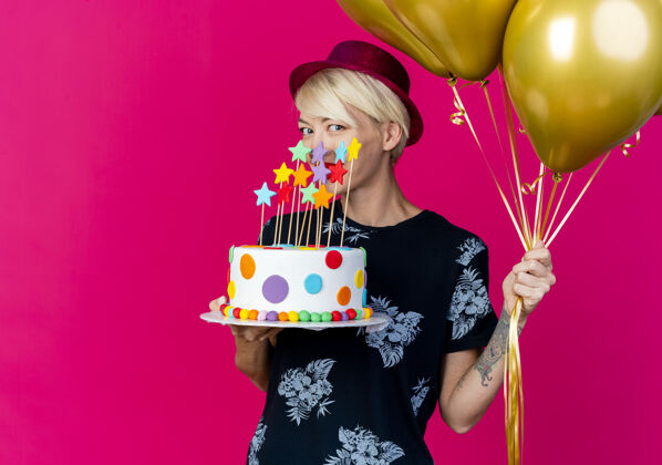 人微笑着的年轻金发派对女郎戴着派对帽拿着气球和生日蛋糕看着前面的星星在粉红色的墙上孤立的复制空间表情脸抱着