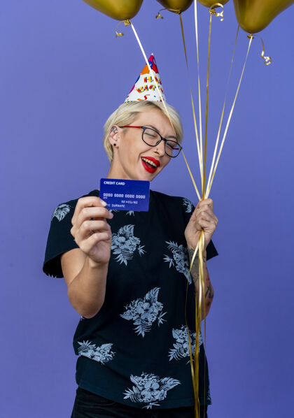 生日快乐的金发女郎戴着眼镜 戴着生日帽 手里拿着气球和信用卡 在紫色的墙前眨眼年轻气球人