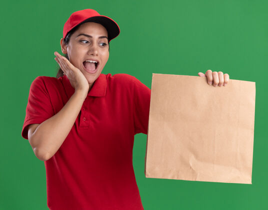 惊喜惊讶的年轻送货女孩穿着制服 戴着帽子 看着纸食品包装把手放在脸颊上隔离在绿色的墙上手帽子年轻