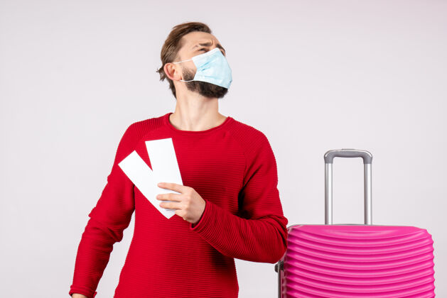 风景正面图年轻男子戴着面具手持机票在白墙上航行covid-航班旅行度假病毒色彩情感机票病毒度假