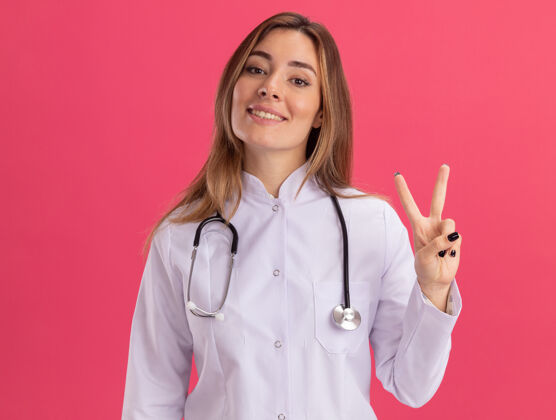穿微笑的年轻女医生穿着医用长袍 听诊器显示出和平的姿态 隔离在粉红色的墙上女性听诊器表演