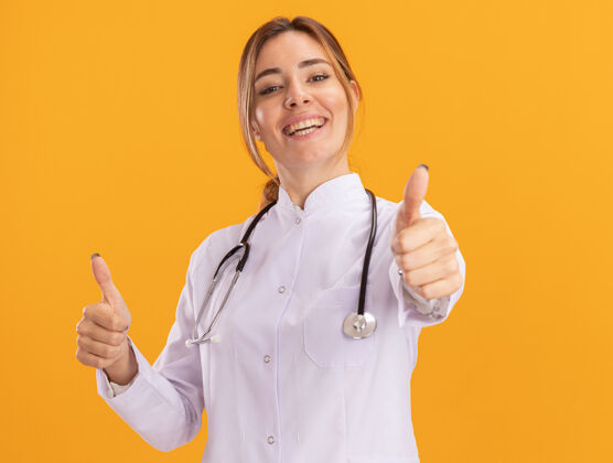 医疗面带微笑的年轻女医生 穿着医用长袍 听诊器竖起大拇指 隔离在黄色的墙上医生表演长袍
