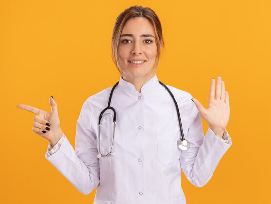 穿高兴的年轻女医生穿着医用长袍 听诊器显示停止手势点隔离在黄色墙上医生女性医疗
