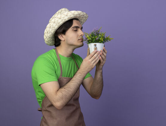 制服高兴的年轻男园丁穿着制服 戴着园艺帽 手持和嗅着紫罗兰上孤立的花盆里的花穿紫色花盆