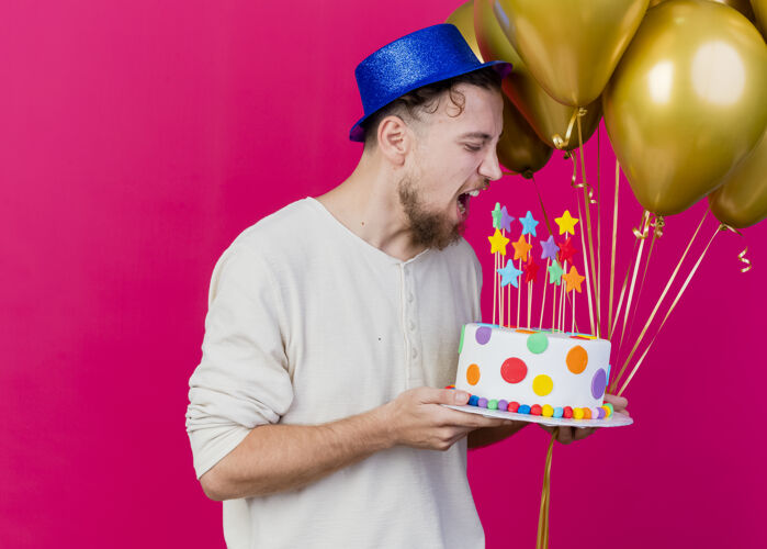 气球年轻帅气的斯拉夫派对男孩戴着派对帽拿着气球和生日蛋糕 明星们正准备咬蛋糕 蛋糕被隔离在粉红色的墙上 还有复制空间明星姿势抱着