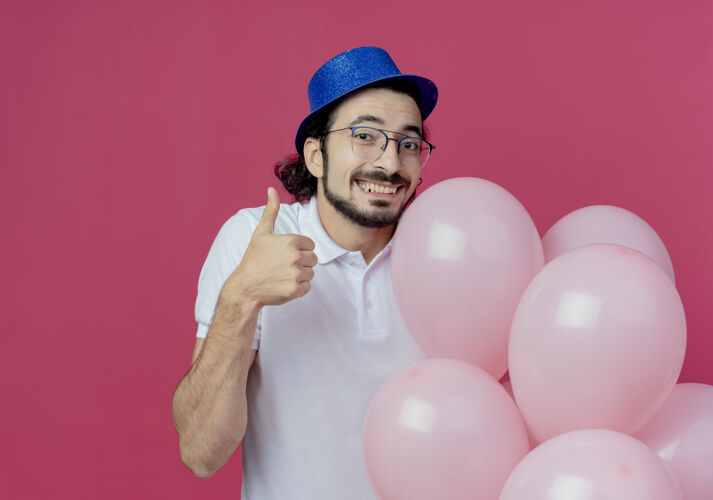 气球笑容可掬的帅哥戴着眼镜 戴着蓝色的帽子 手里拿着气球 大拇指孤立地竖起 背景是粉色的微笑拿着眼镜