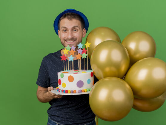 脸微笑的帅哥戴着蓝色的派对帽 手里拿着氦气球和生日蛋糕 隔离在绿色的墙上 还有复制空间氦聚会气球