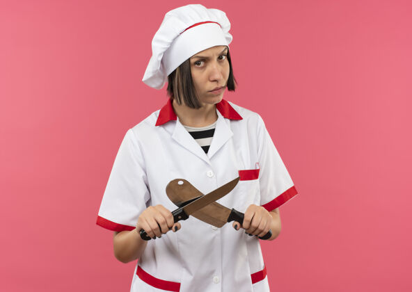 刀穿着厨师制服的年轻女厨师拿着刀和菜刀 粉色背景上有复印空间不愉快制服烹饪